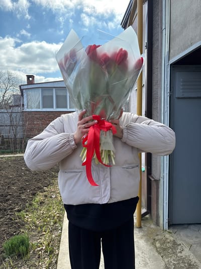 An individual stands outdoors in Trudobelikovskiy, Russia, holding a large bouquet of red tulips, which obscures their face. The person is wearing a light beige puffy jacket, a dark undershirt, and dark trousers. A silver-colored ring is visible on the ring finger of the left hand. The bouquet is wrapped in clear plastic and white paper, tied with a red ribbon. The immediate background to the left shows a patch of tilled earth with emerging green shoots, a wire fence, and a brick building with windows and a chimney. To the right, a light brown building wall, a dark door or gate, and a concrete path are visible. A yellow pipe is partially seen behind the individual. The sky above is blue with scattered white clouds.