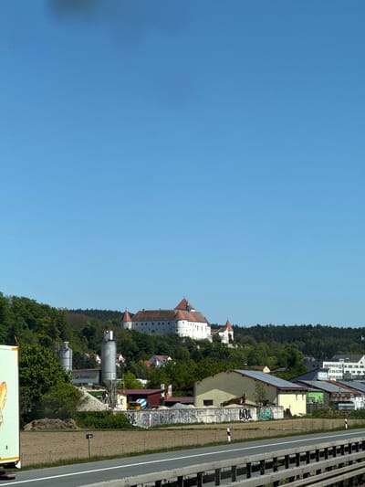 This is an outdoor shot of Wörth an der Donau in Germany. A large white castle with red roofs stands out against the backdrop of a blue sky and green trees. Nearby is an industrial site with large silver silos. A road runs along the edge of the scene, and what appears to be a truck is visible in the foreground. Houses are scattered among the trees, blending into the landscape.