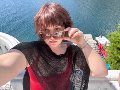 A person with brown hair, sunglasses, a red top, and a black cover-up poses in front of a body of water. A dock with boats and kayaks is visible behind them. They wear a silver bracelet on their left wrist and hold the sunglasses with their hand. A white railing is also visible.
