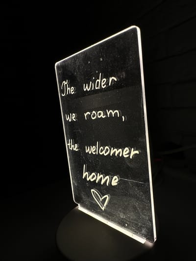 An illuminated sign with white text on a black background is displayed against a dark, possibly indoor, setting. The text, written in a cursive font, reads: "The wider we roam, the welcomer home," followed by a small heart symbol. The sign is transparent and appears to be made of acrylic, with a light source integrated into its base, causing it to glow. The sign is propped up on a dark circular base. The background consists of a dark, textured surface. There are no people or animals present in the image. The scene does not depict any identifiable landmarks or features of Podilsk, Ukraine.