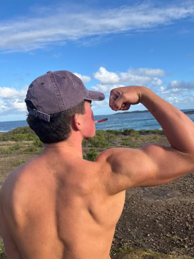 A shirtless man with muscular arms poses against a backdrop of blue sky, clouds, and a body of water. He wears a baseball cap and has a cigarette in his mouth. His right arm is flexed, displaying his bicep, while his back reveals muscle definition. Vegetation and a dirt ground are visible in the background, suggesting an outdoor location.