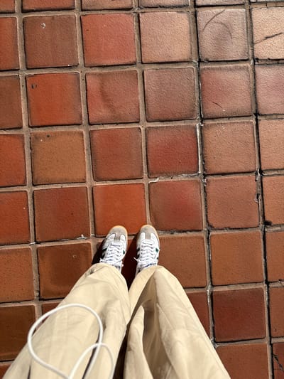 The image presents a high-angle, top-down view of a person's lower legs and feet standing on a tiled surface. The person is wearing light-colored, possibly beige or khaki, baggy pants that gather around the ankles. On their feet are white athletic sneakers with light grey suede accents on the toe and heel, and distinctive dark green stripes along the sides. White laces are tied neatly. A thin white cable, possibly from headphones or a charging device, is visible draped over the right pant leg.

The ground beneath the person is covered in a grid of square, reddish-brown tiles. These tiles vary subtly in color, displaying shades of terracotta, rust, and earthy browns, with visible, darker grout lines between them. Some tiles show minor wear, dust, or slight discoloration. The strong, direct sunlight casts distinct shadows from the person's legs and pants onto the tiles, indicating bright, clear weather, likely during the day. The scene is static, capturing the person in a standing position. There is no visible text in the image. The setting appears to be an outdoor paved area, such as a sidewalk or patio, consistent with environments found in Hialeah, United States of America.