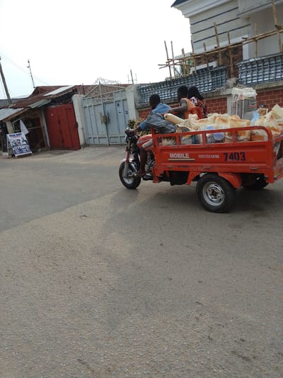 The image captures a motorized tricycle in motion on a paved road. The tricycle is painted red, with the word "MOBILE," a phone number (09123324090), and the number "7403" prominently displayed on its side. The tricycle is loaded with bags, possibly cement, and carries three individuals: a driver and two passengers. The driver is wearing a blue shirt, while the other passengers wear casual clothing.

In the background, buildings and structures are visible. To the left, there is a small, possibly commercial building with a red facade. Adjacent to this building is a sign advertising a barber shop called "NEW BOY." Further in the background, there is a gate and the facade of a larger building, possibly a residence, which is under construction as evidenced by the bamboo scaffolding erected around the building.

The image was captured during daylight hours, under an overcast sky, and the location appears to be Ohokobe Ndume, Nigeria. The scene suggests a typical urban environment in the region, with commercial activity and ongoing construction present.