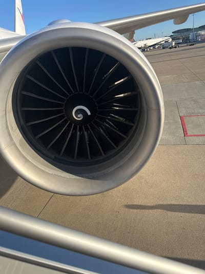 The image, captured in Tremblay-en-France, France, presents a close-up view of the intake of a jet engine mounted on an airplane wing. The engine's metallic, silver-grey cowling, detailed with rivets, dominates the foreground. Inside the circular intake, numerous dark fan blades are visible, along with a prominent white spiral pattern on the central spinner cone.
To the left, a portion of the airplane's wing is seen against a clear, bright blue sky. In the background, another airplane is parked on a light grey concrete tarmac. This aircraft is distinctly branded with "AIR CANADA" visible on its white fuselage and a partial red and blue logo on its tail. Other airport ground equipment and vehicles are faintly discernible near the Air Canada plane. The ground features red painted markings in places. On the right side of the image, a shiny, metallic railing, likely part of an air stair or jet bridge, is partially visible.
The scene is brightly lit by natural sunlight, indicating a clear, sunny day, with shadows suggesting a mid-day to afternoon setting. There are no people visible, and no active events are taking place, depicting a parked aircraft at an airport.