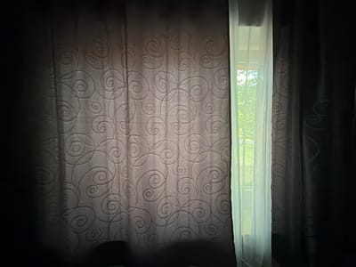 The image depicts a window covered by curtains in Dorsten, Germany. The window is partially obscured by two layers of curtains. The left curtain is a darker, textured fabric with a subtle swirling pattern. To the right of this, a sheer, white curtain is drawn back, revealing a view of the outdoors. Through the sheer curtain, greenery and trees are visible, suggesting it is daytime and likely fair weather. The lighting indicates that the sun is shining, casting a bright glow through the sheer fabric. The overall mood is somewhat subdued due to the contrast between the dark, heavy curtains and the bright light from the window, creating a sense of quiet and perhaps a hint of mystery. There is no visible text in the image.