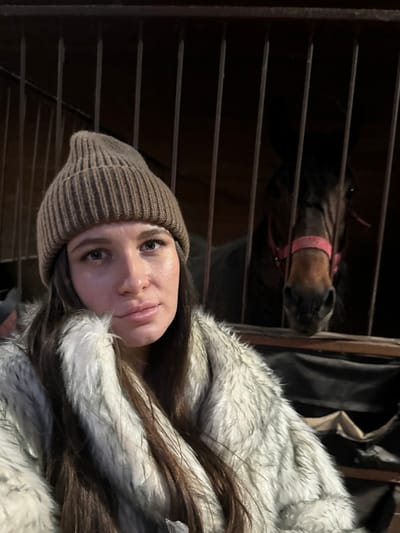 The image depicts a woman and a horse inside a stable, located in Kalinovo, Russia. In the foreground, a woman with long, dark brown hair wears a light brown ribbed beanie and a grey and white faux fur coat. She has a neutral facial expression and is looking directly forward towards the camera. Behind her, separated by dark vertical metal bars, the head and partial neck of a dark brown horse are visible. The horse wears a pink halter and its eyes are directed towards the camera. The background consists of dark wooden stable walls and dark fabric materials.