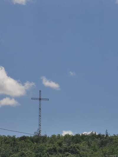 The image depicts a tall, metal cross standing on a densely forested hilltop. The cross is constructed from a lattice of metal beams, forming a geometric pattern. It is silhouetted against a clear blue sky with a few scattered white clouds. Below the hilltop, power lines are visible, strung across the lower part of the frame. The vegetation on the hill is lush and green, suggesting a natural environment. The scene appears to be a landmark or a monument situated on an elevated natural feature, possibly within or on the outskirts of the town of Cărpineni, Moldova. The sky is bright, indicating daytime. There are no people or animals visible in the frame.