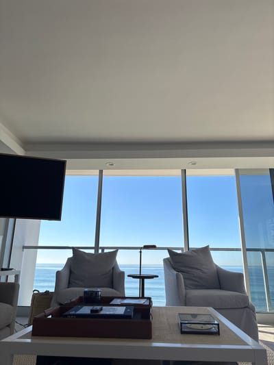 A modern, airy living space features floor-to-ceiling windows offering a stunning ocean view. Two striped armchairs with matching pillows face the vista. A dark TV screen sits against a light-colored wall. A side table displays decorative items. The space is bright, clean, and designed to maximize the ocean view.