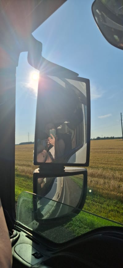 The image is a photograph taken from inside a vehicle, likely a truck, looking out through the side mirrors. The primary subject is a selfie taken by a person holding a smartphone in the upper side mirror. The person appears to be wearing a light-colored top and has their arm raised to take the photo. The reflection in the mirror shows part of the vehicle's interior, including a headrest. The background visible in the mirrors and outside the window is a rural landscape with a field of dry crops under a clear blue sky with bright sunlight and sun flares. A power line is visible in the distance. The lower side mirror reflects the road and the side of the vehicle. The image was captured in or near Kokui, Russia.