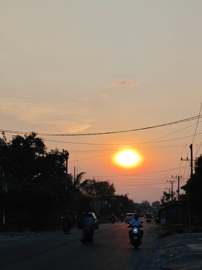 The image depicts a street scene in Blangcot Tunong, Indonesia, at dusk with the sun low on the horizon. A large, intensely bright, circular orange-yellow sun dominates the sky, positioned slightly left of center above the distant tree line. The sky exhibits hues of orange and yellow near the horizon, transitioning to lighter tones higher up, with minimal cloud cover.
A two-lane asphalt road extends from the foreground into the background, where traffic flows in both directions. In the foreground, a motorcycle with a prominent circular LED headlight is ridden by an individual wearing a light-colored, possibly white, collared shirt, facing away from the viewer. Behind this motorcycle, several other vehicles, primarily motorcycles with illuminated headlights and taillights, are visible traveling in both directions, appearing as silhouettes against the setting sun or as bright points of light. A light-colored car is also discernable further down the road on the left, and a dark-colored van-like vehicle is moving away on the right.
The left side of the road is bordered by a dense, dark mass of trees and foliage. Overhead utility poles with multiple crisscrossing wires are visible above the tree line on both sides of the road. On the right side, the environment appears less densely vegetated, with a small, dark structure, possibly a shack or vendor stall, topped with a blue tarpaulin roof, situated near the road. The immediate ground on the right shoulder appears unpaved and somewhat uneven. The overall scene is characterized by the low, warm light of the setting sun, casting significant portions of the foreground and roadside elements into silhouette.