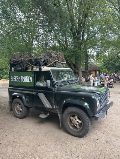 A dark green, rugged Land Rover Defender is parked on a sandy path in an outdoor setting with trees in the background. The vehicle has "BEEKSE BERGEN" emblazoned in white text on its side. There is camouflage netting and what appears to be luggage on the roof rack. A woman is visible in the driver's seat, and another person can be seen in the passenger seat. In the background, a group of people are gathered around picnic tables, with thatched-roof structures and more trees forming the environment. The overall scene suggests a safari or park-like setting in Beekse Bergen, a safari park located in Hilvarenbeek, Netherlands.