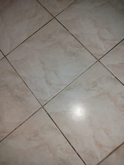The image shows a tiled floor in an indoor location in Bochnia, Poland (latitude: 49.952, longitude: 20.422). The floor is made of beige tiles with a diamond pattern formed by the grout lines. The tiles are slightly worn and dirty with dark spots. The light source is unknown. It's impossible to determine the specific room or building based on the provided image.