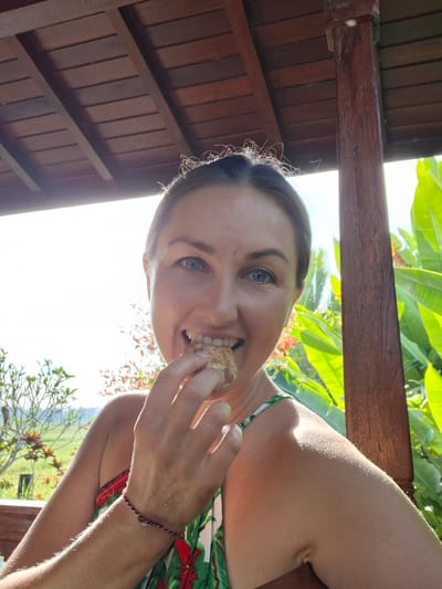 The image is a selfie of a woman eating something while sitting outdoors, likely in a tropical location. The woman has fair skin, light eyes, and her hair is pulled back. She is wearing a top with a green and red leaf pattern. She is holding a round, possibly bread-like food item to her mouth. She appears to be inside a wooden structure with a slatted roof. Outside, there is lush green foliage, including banana leaves and other tropical plants. The location is Lodtunduh, Indonesia.