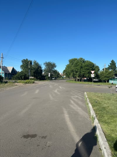 The image depicts a residential street scene in Kalinino, Russia, under a clear blue sky, suggesting a sunny day, likely in the morning or afternoon due to the shadows. The perspective is from ground level, looking down the asphalt road which curves gently to the right. On the left side of the road, a row of lush green trees lines the houses and properties. A prominent feature is a give way traffic sign with a white background and a red triangle. Further down the street, utility poles with power lines are visible, indicating a residential area. On the right side of the road, there is a patch of green grass with a concrete curb. A shadow, potentially cast by a person or an object, falls across the asphalt. The overall impression is a quiet, everyday scene in a rural or suburban setting.
