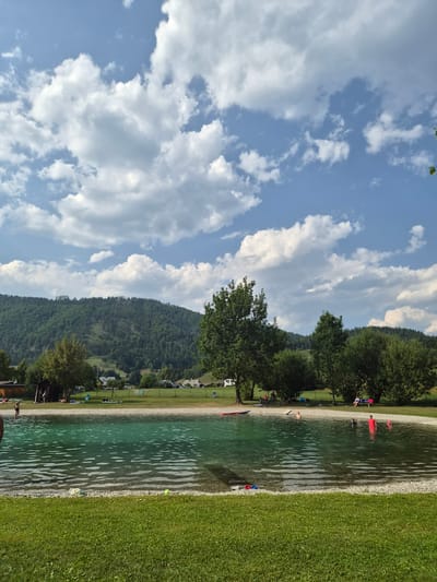 This is a medium shot, eye-level photograph taken on a sunny day at an outdoor swimming area in Landl, Austria. The scene depicts a clear, turquoise water pool surrounded by green grass and a pebble beach. In the background, a dense, tree-covered mountain dominates the landscape, with a few buildings and houses scattered across its lower slopes and in the valley. The sky is a vibrant blue, adorned with large, fluffy white clouds, indicating fair weather.
Several people are visible in and around the pool. In the water, a person in a red swimsuit is standing, while others are swimming or wading. On the pebble beach, a few individuals are relaxing or observing. To the left of the pool, there are two small wooden structures, possibly changing rooms or sheds, with a few people nearby. In the grassy area, lounge chairs and a red inflatable mat are visible. The overall atmosphere is one of recreation and leisure on a warm day.