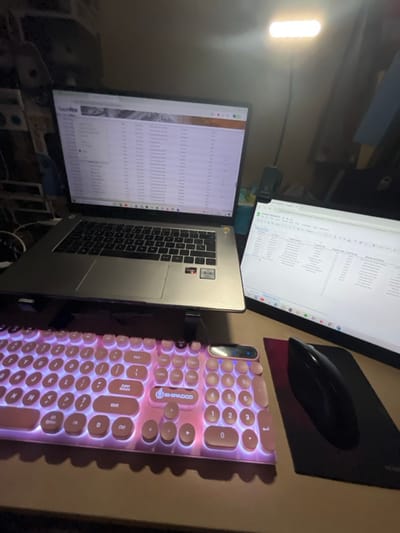 The image depicts an indoor computer workstation in the city of San José Pinula, Guatemala. The scene is dimly lit by ambient light, supplemented by two illuminated screens and a desk lamp.
A silver-grey laptop with a dark keyboard is positioned centrally on a stand, its screen displaying a web browser window titled "CoolView" with tabular data, likely a list or database entries. An AMD Ryzen sticker is visible on the laptop's palm rest. To the right of the laptop, an external flat-panel monitor is oriented vertically, displaying a spreadsheet application with multiple columns of data including headers like "Product ID" and "Property Type."
A backlit keyboard with circular, pink keycaps, emitting a pink/purple glow, is positioned directly in front of the laptop. The brand "SHIPADCO" is visible on the spacebar. To the right of the keyboard, a black optical mouse rests on a black mousepad.
A flexible-neck LED desk lamp, featuring a rectangular light panel, is clamped to the right side of the desk, emitting a bright white light that illuminates the external monitor and adjacent desk area. The equipment rests on a light brown desk surface. The background is dark and out of focus, with indistinguishable objects vaguely visible on the left and a hint of a light blue object on the far right. No individuals are present in the frame.
