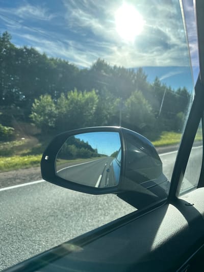 The image captures a view from inside a moving vehicle, looking out the side window and into the side-view mirror. The setting appears to be a rural road with a dense line of green trees bordering the right side of the road. The sky is blue with scattered white clouds, and the sun is shining brightly. The side-view mirror reflects the road ahead, which is asphalt with white lane markings. The interior of the car is visible in the foreground, specifically the door panel and the edge of the window frame. The location is identified as Bol'shoe Ondrovo, Russia. There are no people or direct interactions depicted. The scene is one of travel on a sunny day through a natural landscape.