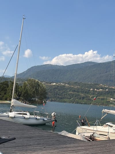 The image depicts a serene waterfront scene at Calceranica al Lago, Italy. The primary subjects are several sailboats moored along a wooden dock. The boats are white with some visible blue accents and nautical equipment such as fenders and masts. One sailboat in the foreground is mostly visible, showcasing its hull and a partially unfurled sail. In the background, a lush green mountainside rises steeply from the lake, dotted with buildings and trees. The water is calm, reflecting the blue sky and the surrounding landscape.
The scene appears to be set during the daytime, with bright sunlight casting shadows and illuminating the wooden planks of the dock. The weather seems pleasant, indicated by the clear blue sky and the presence of scattered, fluffy white clouds. A light breeze might be inferred by the slight ripple on the water's surface. The Italian flag is visible on one of the boats, adding a specific location cue. The overall mood is tranquil and peaceful, suggesting a relaxed atmosphere at the lake.
No people are clearly visible in the image, though there may be individuals on the boats or in the distance that are too small to discern. A piece of text is visible on the side of one of the sailboats, reading "217 S FIRE". The image is oriented vertically, so the scene is viewed from a tilted perspective.