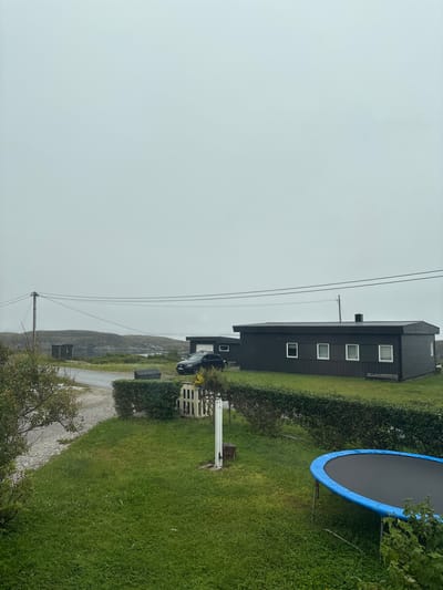 The image depicts a rural setting in Hamarvik, Norway, on an overcast day. A dark, modern house with multiple windows stands prominently on a grassy slope, bordered by a neatly trimmed hedge. Power lines stretch across the frame, and a black car is parked near the house. To the right, a large trampoline with a blue rim and black mat is set up on the lawn. The landscape beyond the house shows rolling hills or mountains, suggesting a coastal or fjord environment. The overall atmosphere is subdued due to the cloudy weather. There are no people visible in the image.