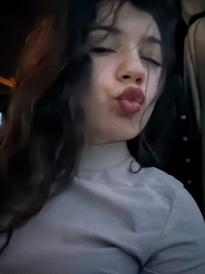 A woman with closed eyes and puckered lips lies in a relaxed pose. Her dark hair is partially visible. She wears a light-colored, possibly grey, long-sleeved shirt. The background is dark and somewhat indistinct, suggesting the photo was taken in low light. The overall impression is one of peacefulness or sleep.
