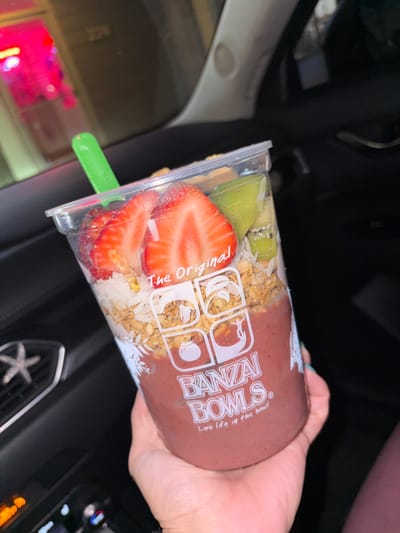 A person holds a "Banzai Bowls" cup filled with an acai bowl, featuring layers of granola, sliced strawberries, and kiwi. The cup has a green spoon inserted. The cup is being held inside a vehicle, given the presence of car interior elements like a black dashboard and seating. Outside the car window, reflected in the shiny plastic of the cup, a nighttime urban environment is visible with the neon lights of business front. Given the branding of Banzai Bowls, there is a strong probability that the location is near Huntington Beach in California where it originated.