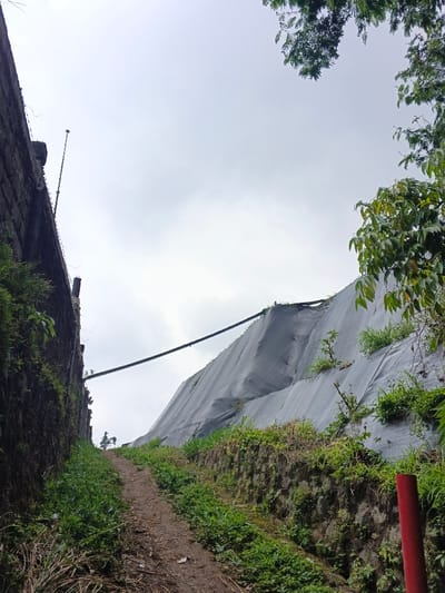 The image displays a narrow, upward-sloping dirt path in Uato Carabar, Timor-Leste, situated between two elevated structures under an overcast sky. To the left, a dark, weathered stone or concrete retaining wall, partially covered in green vegetation and moss, rises steeply. A slender metal pole extends vertically from the top of this wall, and a pipe-like object is visible lower down. To the right, a steep embankment is covered with a dark grey or black erosion control fabric (geotextile), through which patches of green vegetation are growing. Below this fabric-covered slope, a lower stone or concrete retaining wall, also vegetated, defines the path's edge. A thick, dark cable or pipe spans horizontally across the middle ground, connecting the top of the left retaining wall to the upper section of the right embankment. The dirt path is light brown and uneven, flanked by green grass and weeds. A red pole is partially visible in the lower right corner. Green tree foliage is present in the upper left and upper right corners of the frame. No individuals or animals are present.