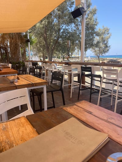 This image captures an outdoor seating area at a beach bar in Protaras, Cyprus. The scene is set under a large, orange awning providing shade. In the foreground, several wooden tables are visible, some with white chairs and others with black high chairs arranged around them. One table has a metal ashtray with the number "51" embossed on it. Further back, a long bar counter is lined with white and black bar stools. Beyond the seating area, trees with light green foliage line a pathway leading towards the sea. The background reveals a clear blue sky and the turquoise waters of the Mediterranean Sea. A few people are present in the distance, walking along the beach. The overall impression is of a relaxed, sun-drenched beachside establishment.