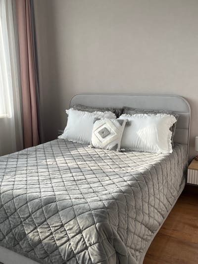The image shows a bedroom scene with a bed in the center. The bed is covered with a gray quilted blanket and has several pillows. Two white pillows with ruffled edges are placed at the back, and a decorative square pillow with a diamond pattern and tassels is in front of them. The headboard of the bed is upholstered in gray fabric. To the left of the bed, there are sheer white and pinkish-gray curtains covering a window, through which sunlight is entering, casting shadows on the bed. The wall behind the bed is a light, neutral color. To the right of the bed, a small nightstand with a wooden top and white ribbed drawers is visible. The floor is made of wooden planks. There is no indication of people, actions, or interactions. The scene depicts a quiet, neatly arranged bedroom. The location is specified as Meleuz, Russia, but this information is not visually verifiable from the image itself.