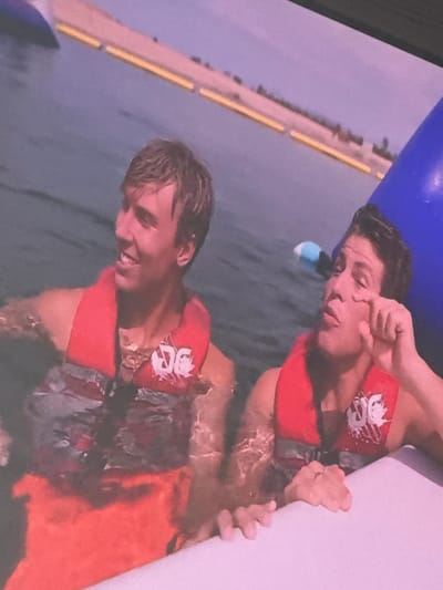 The image shows two young men in life vests in the water. The man on the left is smiling and looking to the left. The man on the right is looking forward and touching his eye. They are in a body of water with a floating structure in the background. The water appears to be from a lake or river. The lighting suggests it is daytime. The scene is outdoors. There are no identifiable landmarks in the image that would allow for precise localization to the city of Moelv, Norway.