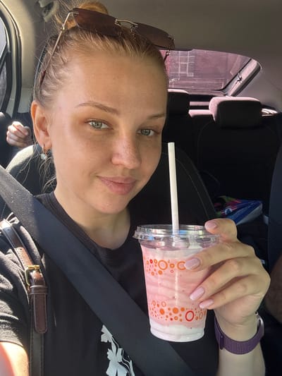 A young Caucasian woman with blonde hair pulled back in a bun and sunglasses perched on her head is seen from the chest up, sitting in the passenger seat of a car. She is wearing a black t-shirt and a seatbelt. She is holding a pink and white milkshake in a clear plastic cup with a white straw. The woman has green eyes, light skin with freckles, and is wearing small hoop earrings. Her nails are painted a light pink. In the background, car seats and a tinted window are visible. There is also a partial view of another person's hand in the upper left of the image. The scene appears to be a sunny day, suggesting an outdoor location. There is no information in the image to suggest the location is Fedorino, Russia.