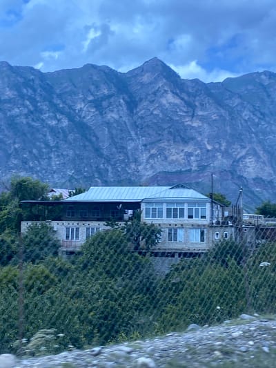 The image shows a stone house with a metal roof situated on a hillside in the village of Irganay, Russia. The house has a balcony and large windows, and is surrounded by lush green trees. In the background, a majestic mountain range looms under a cloudy sky. The scene is partially obscured by a wire fence in the foreground, with rocks and gravel on the ground.