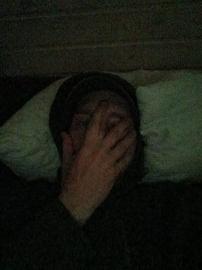 The image shows a person lying on a pillow, with their face obscured by their hand. The person is wearing a dark-colored hat and shirt. The background is dark and appears to be a wall or a blanket. The image is grainy and appears to have been taken in low light conditions. It is impossible to determine the specific location in Zarubino, Russia from the provided image.