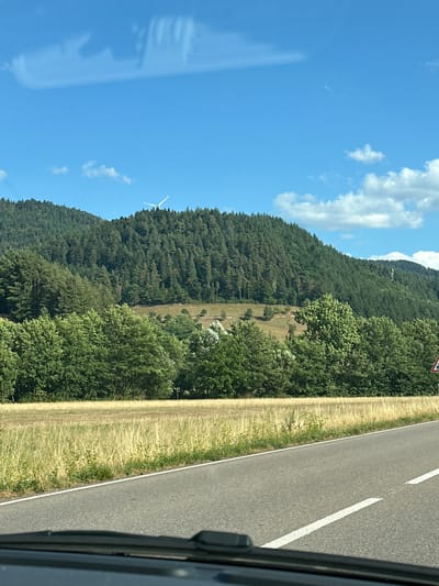The image shows a scenic view of a rural area with rolling hills covered in dense green forest. A clear blue sky with scattered white clouds dominates the upper portion of the frame. In the distance, atop one of the forested hills, a white wind turbine is visible. In the foreground, a paved road runs horizontally across the image, bordered by a dry, grassy field. The perspective suggests the photo was taken from a moving vehicle, as the lower part of the image shows a portion of a car's dashboard. There are no people or animals visible in the scene. The landscape is characteristic of the Black Forest region, likely in or near Gutach (Schwarzwaldbahn), Germany, given the presence of forested hills and a wind turbine, common features in this area.