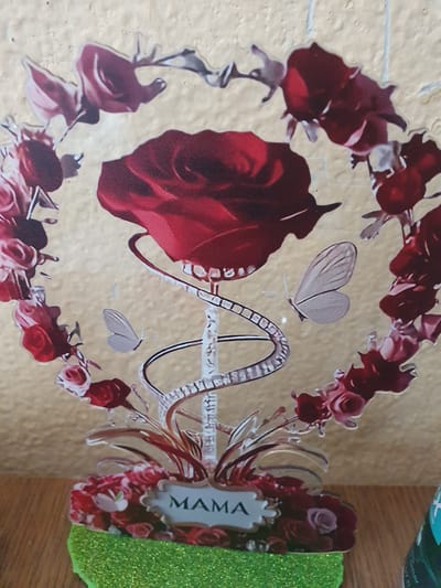 The image shows a close-up of a decorative item featuring a large red rose at the top, surrounded by a heart-shaped wreath of smaller red roses and green leaves. A double helix medical symbol with a silver, jeweled appearance is intertwined with the rose stem. Two small, light-colored butterflies are positioned on either side of the medical symbol. At the base of the decoration, a cluster of red roses and green foliage is visible, with a sign that reads "MAMA" in a decorative font. The entire piece is mounted on a bright green, glittery base. The background is a textured, light tan or beige wall. The scene appears to be an indoor setting, possibly a shelf or table, given the wooden surface visible at the bottom edge of the frame. There is no indication of the city of Dubossary, Moldova, in the image.