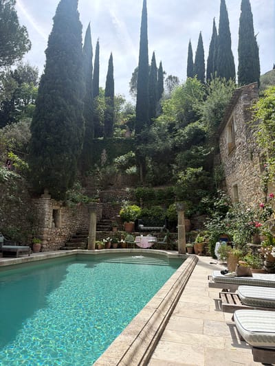 This media file presents a serene outdoor scene, likely a private garden or villa in Saint-Victor-la-Coste, France, captured during daylight hours. The image is rotated 90 degrees, displaying a tranquil setting with a swimming pool, stone architecture, and abundant greenery.

In the foreground, a long, rectangular swimming pool features clear, turquoise water. The water's surface is illuminated by bright sunlight, creating vibrant, rippling patterns of light and shadow. The pool is bordered by light-colored stone coping. Adjacent to the pool, on a paved stone terrace, several wooden lounge chairs with striped, light-colored cushions are arranged, suggesting an area for relaxation.

Beyond the pool, the scene rises in terraces of traditional stone architecture. These robust stone walls and structures, some partially covered with lush green ivy and other climbing plants, form the backdrop. Among the greenery, hints of flowering plants with pink blossoms are visible near the stone walls. In the middle ground, a small table draped with a pink tablecloth and chairs can be seen near a prominent stone pillar.

The background is characterized by numerous tall, slender cypress trees, a hallmark of the Mediterranean landscape, standing vertically against a light sky with a few faint wispy clouds. Various other green trees and shrubs are interspersed throughout the terraced landscape.

No people are visible in the scene, and there is no discernible text. The bright, clear lighting indicates it is daytime with fair weather, contributing to the overall peaceful and idyllic atmosphere of the well-maintained outdoor space.