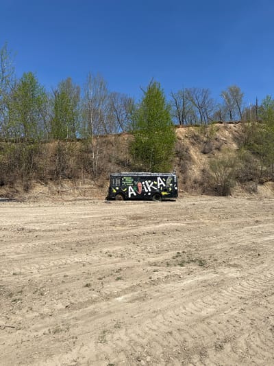 The scene is a dirt lot in Chigiri, Russia, with a black vehicle parked on the right side of the image. The vehicle has graffiti with the word "AIKKA" prominently displayed on its side. The lot shows tire tracks, indicating previous vehicular activity. Behind the vehicle is a steep, vegetation-covered slope. The sky is clear and blue.