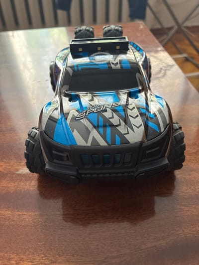 The image shows a close-up, top-down view of a toy remote-controlled car with a blue and silver geometric pattern on its body. The car is black with blue and silver accents. It has large, rugged tires and what appears to be a spoiler with two small lights on top. The car is positioned on a wooden surface, likely a table. The background is blurred, showing vertical lines that could be part of furniture or a wall. There are no people visible in the image. The scene is indoors. The location of Melitopol, Ukraine, is not determinable from the image.