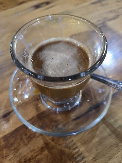 A close-up, high-angle shot shows a glass coffee cup and saucer on a wooden table. The cup is clear with a wide mouth, narrowing towards the base. It contains a light brown liquid with a foamy surface. The saucer is also clear glass, reflecting light and the table's wood grain. A portion of a silver spoon is visible on the right side of the saucer. The background is a blurred wooden surface. The location is Cempeudak, Indonesia.