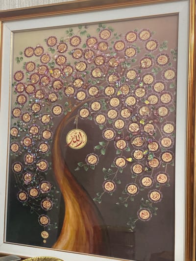 The image shows a framed artwork depicting an Islamic calligraphy tree. The tree has a brown trunk and branches adorned with circular elements, each containing Arabic script, presumably names of Allah or verses from the Quran. These circular elements are surrounded by small purple floral shapes and embellished with translucent, colorful beads. A larger circular pendant with prominent Arabic calligraphy, appearing to be the word "Allah", hangs from a chain in the center of the tree. The background is a gradient of brown and darker tones, with subtle floral patterns. The frame is a double border, with an inner white mat and an outer gold frame. The artwork is displayed indoors, against a textured light-colored wall. There are no people visible in the image. The artwork's placement in Zhanaozen, Kazakhstan is not indicated by any visual cues within the image itself.