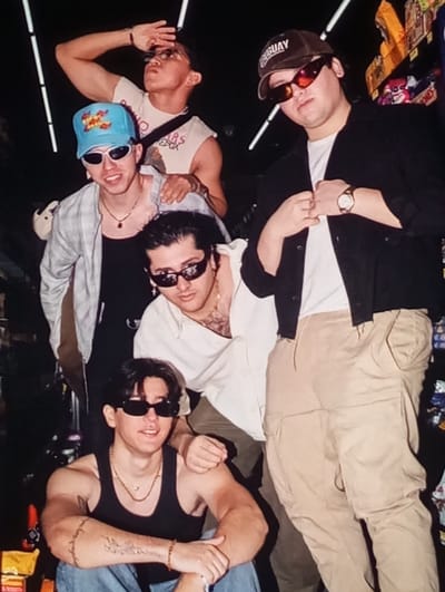 This image displays five males posing in a commercial interior located in Kuyi Charhin, Uzbekistan. The background features dimly lit retail shelving stocked with various products and overhead fluorescent tube lighting.
The individuals are arranged in a staggered group configuration:
1. One male is positioned elevated at the top-center-back, wearing clear-framed glasses and a light-colored sleeveless top. His right hand is raised to shield his eyes, and his head is tilted back.
2. To his left and slightly below, a male wears a light blue cap with colorful embroidery, small white oval sunglasses, a black tank top, and an unbuttoned light-colored long-sleeved shirt. A dark pendant necklace is visible. His left hand rests on the shoulder of the male positioned below him.
3. In the middle-center, a male with dark hair wears dark rectangular sunglasses, earrings, and an unbuttoned light-colored long-sleeved shirt, revealing chest hair and a tattoo. His right hand rests on the shoulder of the male seated in the foreground.
4. At the bottom-front, a male is seated, wearing dark rectangular sunglasses, a black tank top revealing muscular arms with tattoos (a barbed wire design on the upper left arm, text on the forearm), bracelets, and light blue jeans. A gold chain necklace is visible. His hands are clasped in front of him.
5. On the right side, standing, a male wears a dark brown baseball cap with "URUGUAY" lettering, large dark-orange tinted sunglasses, a white t-shirt under a black unbuttoned long-sleeved shirt, and light brown cargo pants. A gold watch is visible on his left wrist, and his hands are positioned near his chest.
All subjects are looking generally towards the camera with neutral to slightly smiling expressions. The scene is illuminated by artificial lighting, highlighting the foreground subjects.