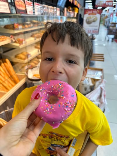 A young boy is being fed a pink donut with sprinkles by a hand. He is wearing a yellow t-shirt and appears to be in a bakery. The bakery has shelves of bread and pastries visible in the background, as well as signage in Vietnamese. The boy's face is partially obscured by the donut he is eating. The hand holding the donut has red painted fingernails. The overall impression is a candid moment of a child enjoying a treat.