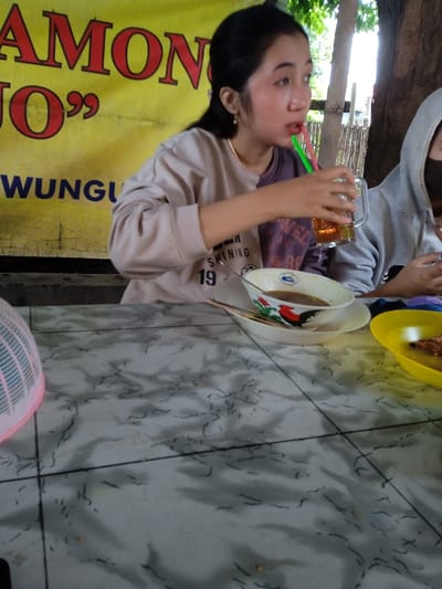 A young woman sips from a glass with a green straw, seated at a table with a bowl of soup with a chicken design. A second person in a hooded sweatshirt sits to her right. A yellow banner with black lettering is partially visible behind her. The table is marble-patterned and has a yellow plate with food to the right of the bowl and a pink plastic basket in the lower-left corner.