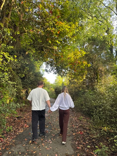 A man and a woman are walking hand-in-hand on a path through a dense forest. The woman is wearing a white blouse and brown pants, while the man is wearing a white shirt and black pants. The path is covered with leaves. The location is Tsanbaikwara, Abkhazia, as inferred.