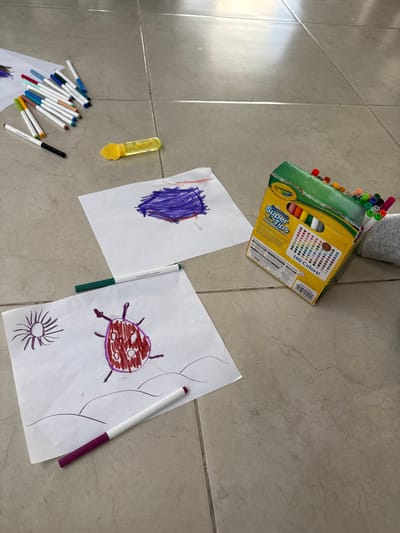 The image displays an indoor scene on a light-colored tiled floor in Aguascalientes City, Mexico. Several art supplies and finished drawings are scattered across the floor. Three pieces of white paper are visible: one in the lower-left corner depicts a red and purple insect-like creature with a purple sun and black ground lines; another in the upper-right features a purple abstract drawing over faint orange lines; and a third, partially visible sheet in the upper-left, shows blue markings. A collection of assorted colored felt-tip markers lies dispersed on the floor, alongside a yellow, transparent plastic object, possibly a glitter container. A partially crushed "Crayola Super Tips 100 Colors" cardboard box stands upright on the right, filled with numerous markers. A lower leg, clad in a light grey sock, is partially visible on the far right of the frame.