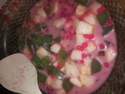 This image displays a close-up, overhead view of a mixed dessert or beverage contained within a translucent bowl, located in Doramamidi, India. The mixture consists of a pink, opaque liquid, irregularly shaped white translucent chunks, similarly shaped translucent green gelatinous pieces, and numerous small, spherical, opaque red elements distributed throughout. A white plastic slotted spoon, stained with pink residue, is partially visible in the bottom-left portion of the frame, resting within or beside the bowl. The bowl's rim exhibits a textured, possibly ribbed or woven pattern. The background is dark, providing no additional environmental context.