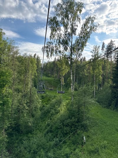 The image depicts a panoramic view from a ski lift in the city of Svetloe, Russia, looking down a cleared slope through a dense forest during a verdant, non-winter season. The upper portion of the frame features a blue sky with scattered, horizontally elongated white clouds. A prominent dark cable, part of the ski lift system, runs diagonally from the upper left to the middle right. Multiple empty, metallic-grey ski lift chairs are visible along the cable, receding into the distance over the green slope. The cleared path, likely a ski run, is covered with lush green grass and dense undergrowth. On both sides of the path, the landscape is dominated by a thick forest composed of various tree species, including several tall birch trees with white bark and numerous other deciduous and coniferous trees with dense green foliage. In the lower right quadrant, a small, light-colored, rectangular object, possibly a sign, is partially visible leaning against the ground vegetation. No individuals are present in the scene.