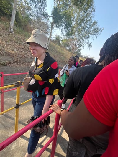 The image depicts an outdoor scene in Kunkwaba, Nigeria, under bright daylight. A group of individuals is gathered on a paved pathway, bordered by a multi-colored barrier featuring red horizontal rails and vertical yellow and red posts.
In the left foreground, a fair-skinned individual, possibly exhibiting albinism, walks towards the right. They wear a light tan bucket hat with a dark "GUCCI" logo, a black short-sleeved button-up blouse adorned with large red, yellow, white, and blue polka dots, and dark blue jeans. A light brown quilted shoulder bag with a gold chain strap hangs on their left side, and they hold a dark, striped object in their left hand near the railing. Gold hoop earrings are visible on their ears.
To the immediate right, partially obstructing the view, is the shoulder and arm of a dark-skinned person wearing a red short-sleeved shirt. Their right hand rests on the red railing. Behind them, another dark-skinned individual wears a black t-shirt, grey jeans, and white sneakers, also near the railing. Further back, a dark-skinned woman in a light-colored striped shirt and dark pants stands holding a green mesh bag and a smartphone. Several other dark-skinned individuals are visible in the background, standing in what appears to be a queue or group.
The background features an upward sloping embankment covered with dry, light-colored vegetation and several trees with light bark and sparse green leaves. The sky is clear and bright blue. Strong sunlight casts distinct shadows on the ground.