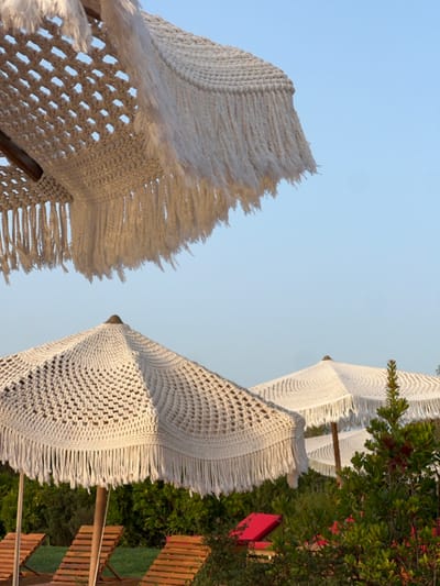 The image depicts an outdoor leisure area in Aljezur, Portugal. The scene is dominated by multiple cream-colored, crocheted sun umbrellas, each featuring an open-weave pattern and a fringed edge. One umbrella is partially visible in the upper-left frame, showcasing its intricate underside. A second, fully visible umbrella stands in the lower-left, supported by a wooden pole, with its pattern and fringe clearly discernible. Two additional similar umbrellas are visible in the background to the right. Beneath the umbrellas, several slatted wooden sun loungers are present, and a red lounge cushion or chair is partially visible amidst dense green foliage in the mid-ground. The sky above is clear and light blue. No individuals are present in the scene.
