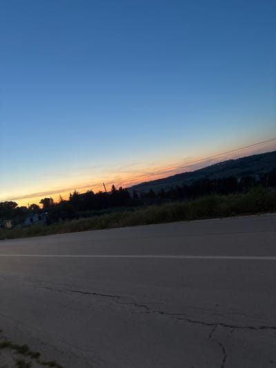 The image shows a roadside scene at twilight in Jesi, Italy. The sky transitions from deep blue to orange near the horizon, suggesting either sunset or sunrise. A dark treeline and vegetation run along the edge of a paved road with a white lane marking. The road surface is visibly cracked and worn. The view is angled, making it difficult to discern specific buildings or other landmarks, but the presence of vegetation and a road suggests a semi-rural or outskirts location of Jesi.