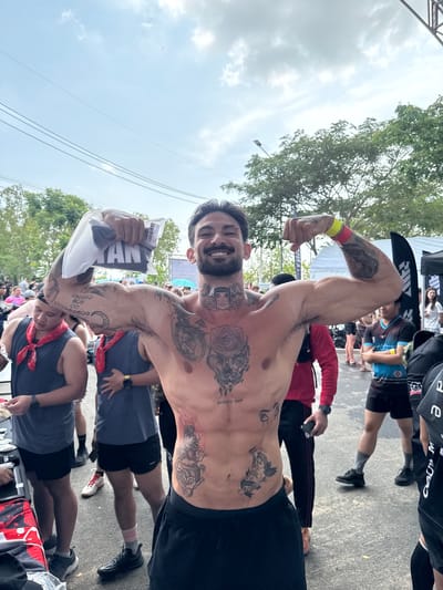 The image shows a shirtless man with multiple tattoos flexing his arms, likely at a race or event. He is holding a packaged item with the word "MAN" visible on it. The setting is likely at the finish line of the race in the city of บ้านสำนักตะแบก, Thailand, due to the presence of other participants and event-related structures nearby. The man is surrounded by other runners, some wearing tank tops with red bandanas. The weather appears overcast.